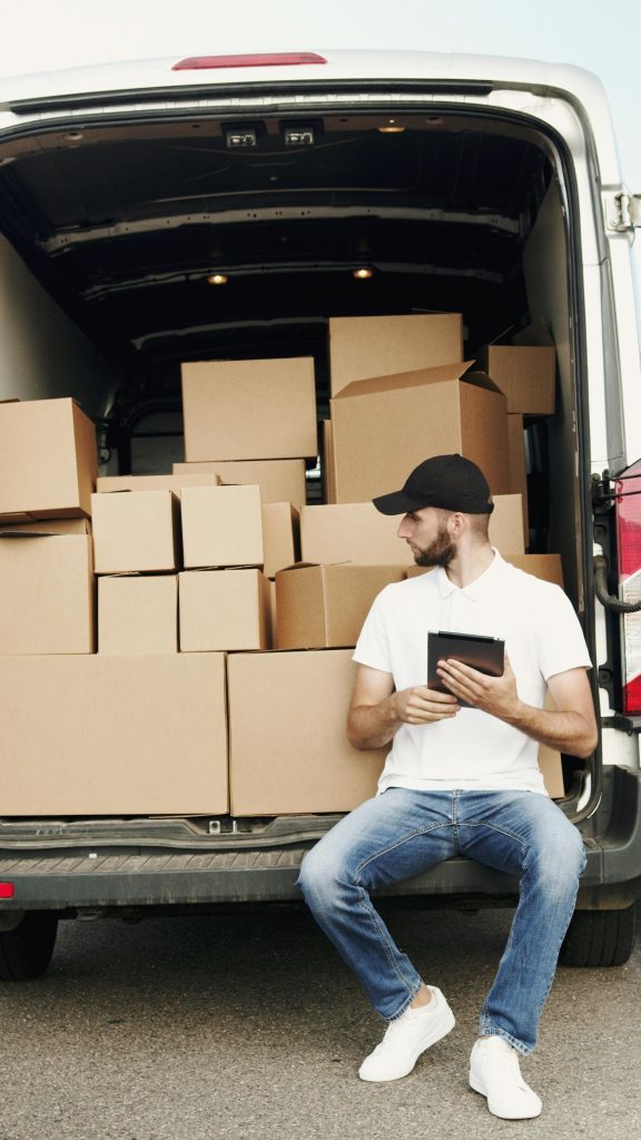 A courier sitting by a van filled with packages, using a digital tablet.