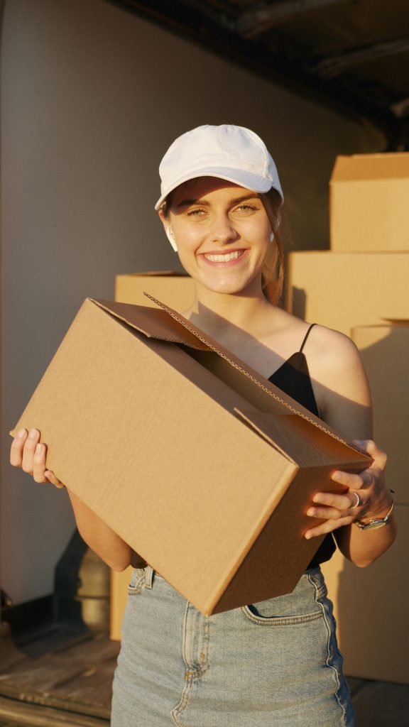 A cheerful woman wearing a cap holds a cardboard box with sunlight illuminating her face.